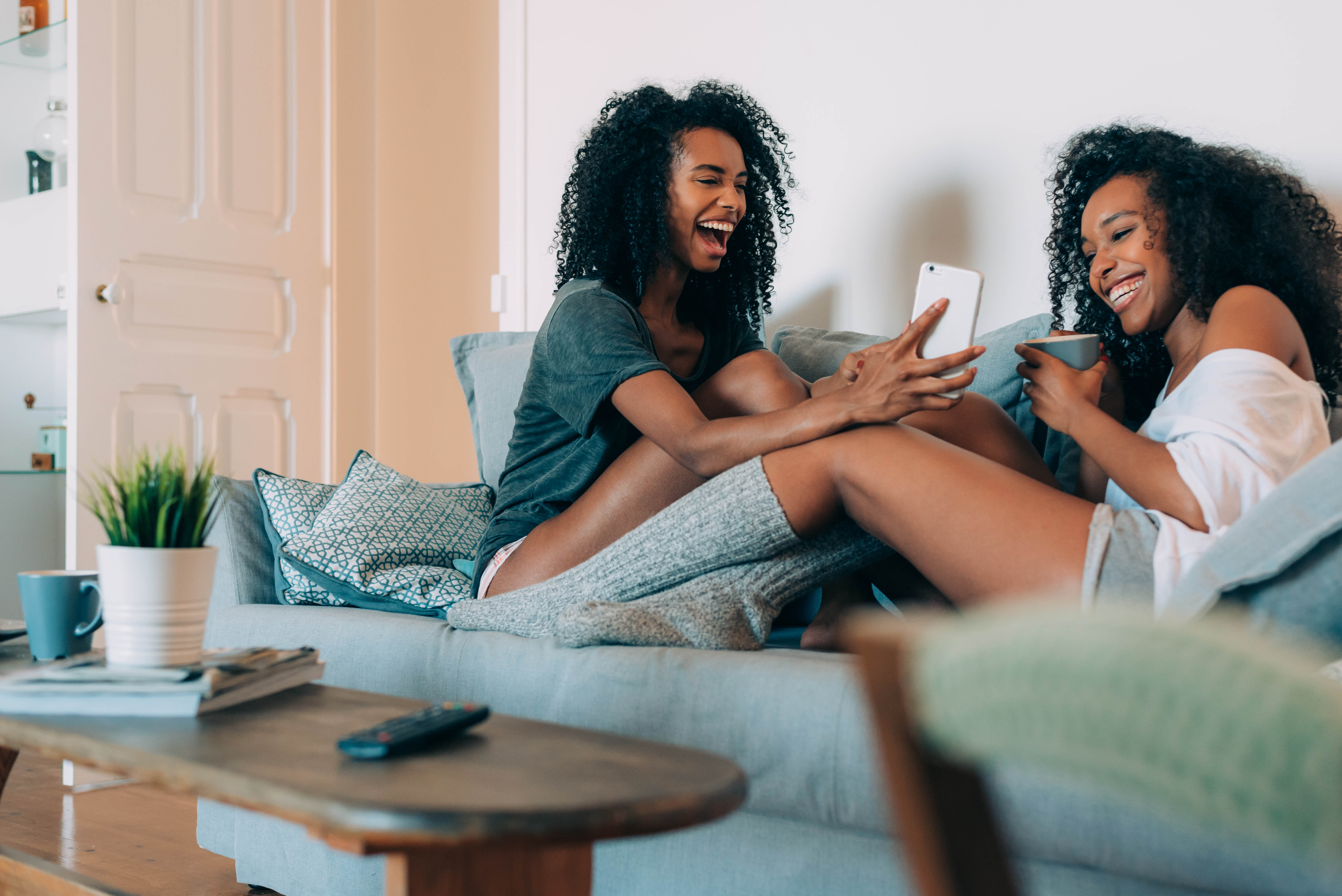 Two young black women sitting on a couch with their phones, laughing and talking Two young black women sitting on a couch with their phones, laughing and talking